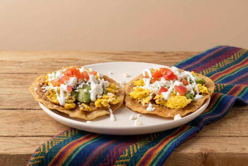 Two breakfast tostadas with scrambled eggs, avocado, tomatoes, cheese, and crema on a colorful napkin.